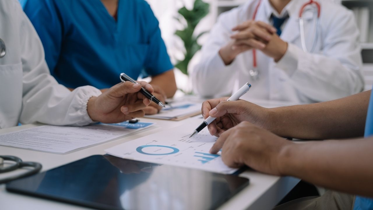 A close-up, eye-level shot shows four healthcare professionals in white lab coats and blue scrubs gathered around a white table. Their faces are out of frame. They are reviewing medical documents and charts, with two people pointing at a donut chart and bar graph using pens.