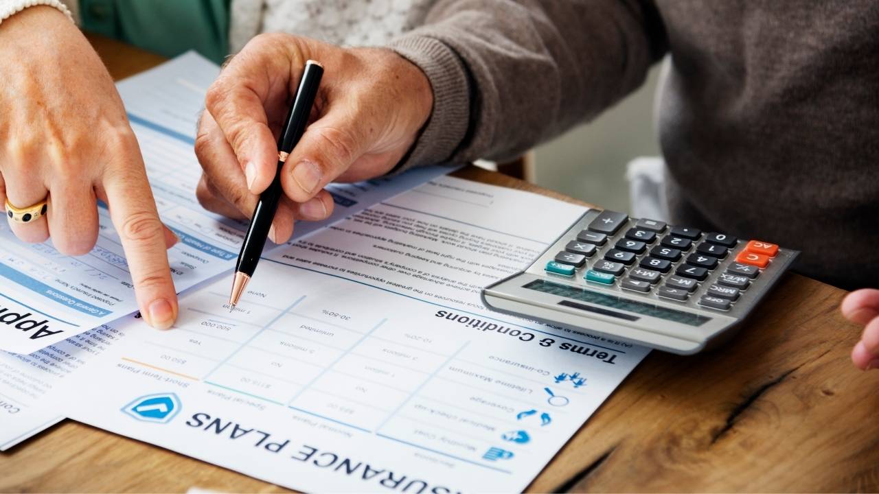 Blockchain in Healthcare, Moving Toward Frictionless Claims A close-up, high-angle shot of two people reviewing a paper document titled "INSURANCE PLANS" on a wooden table. One person points a finger at a section of the document, while another holds a black and gold pen over the page.